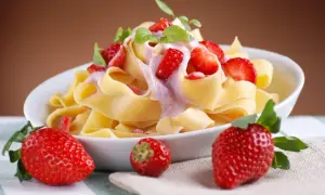 Pappardelle z kremowym sosem i kawałkami świeżych truskawek, ozdobione listkami mięty. Obok talerza leżą dojrzałe truskawki.