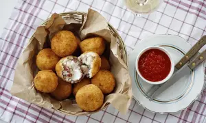 Koszyk pełen złocistych arancini, czyli smażonych kulek ryżowych z serem i mięsem. Obok miseczka sosu pomidorowego.