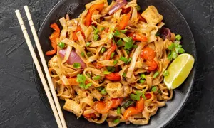 Pyszny pad thai przepis: makaron ryżowy z tofu, papryką, cebulą i groszkiem, udekorowany kolendrą i limonką.