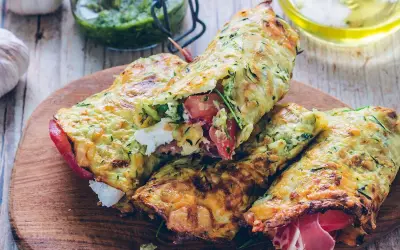 Złociste tortilla z cukinii, nadziewane pomidorami, fetą i szynką, prezentowane na drewnianej desce.