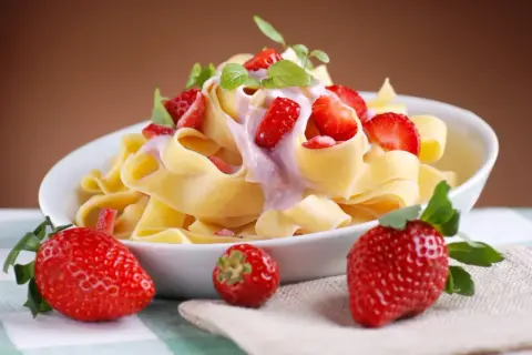 Pappardelle z kremowym sosem i kawałkami świeżych truskawek, ozdobione listkami mięty. Obok talerza leżą dojrzałe truskawki.