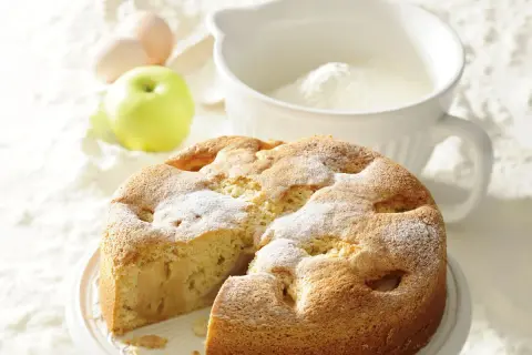 Puszyste ciasto ucierane z jabłkami, posypane cukrem pudrem, obok jabłko, jajka i miska z mąką.