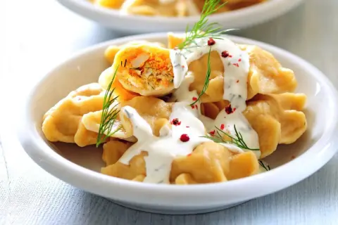 Pyszne, szybkie dania bezmięsne na obiad: pierogi z farszem warzywnym, polane sosem jogurtowym z koperkiem i posypane pieprzem.