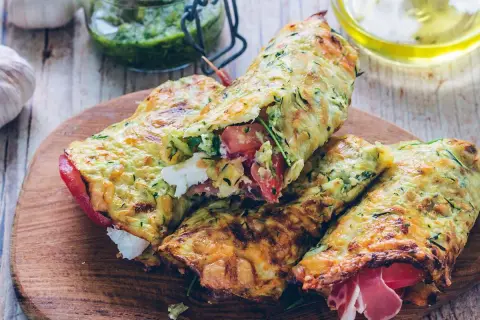 Złociste tortilla z cukinii, nadziewane pomidorami, fetą i szynką, prezentowane na drewnianej desce.