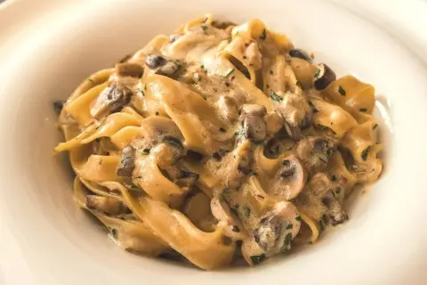 Pappardelle z kremowym sosem i kawałkami pieczarek, posypane natką pietruszki. Pyszny makaron z pieczarkami w białym talerzu.
