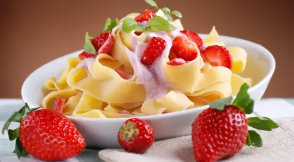 Pappardelle z kremowym sosem i kawałkami świeżych truskawek, ozdobione listkami mięty. Obok talerza leżą dojrzałe truskawki.