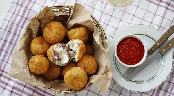 Koszyk pełen złocistych arancini, czyli smażonych kulek ryżowych z serem i mięsem. Obok miseczka sosu pomidorowego.