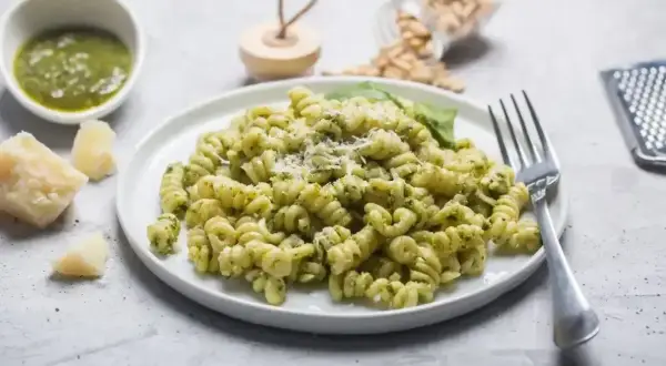 Pyszny makaron z pesto, posypany parmezanem, obok kawałki sera i miseczka sosu. Idealny posiłek dla miłośników włoskich smaków.