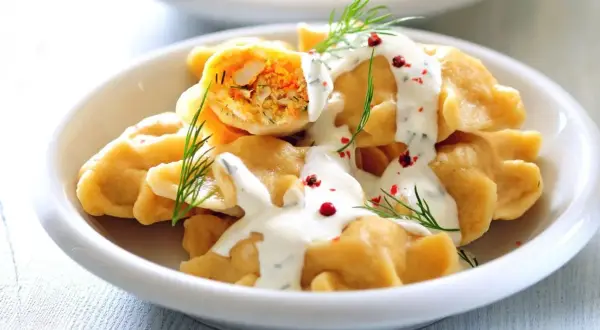Pyszne, szybkie dania bezmięsne na obiad: pierogi z farszem warzywnym, polane sosem jogurtowym z koperkiem i posypane pieprzem.