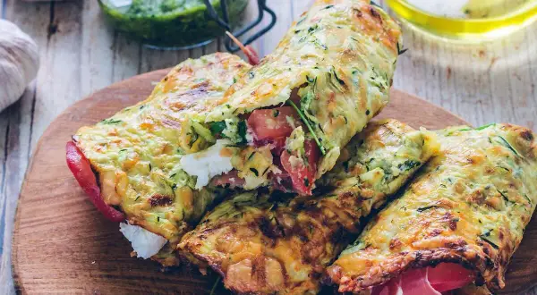 Złociste tortilla z cukinii, nadziewane pomidorami, fetą i szynką, prezentowane na drewnianej desce.