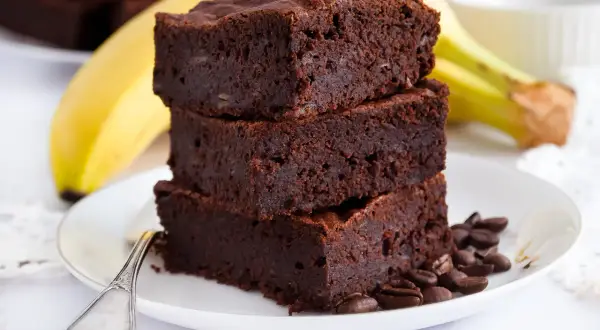 Stos puszystych brownie z bananami, ozdobiony ziarnami kawy, na talerzu z bananem w tle.