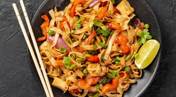 Pyszny pad thai przepis: makaron ryżowy z tofu, papryką, cebulą i groszkiem, udekorowany kolendrą i limonką.