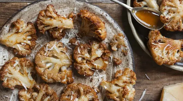 Pieczone dania z kalafiora, posypane startym parmezanem, na rustykalnym talerzu. Obok łyżka sosu i kawałek sera.