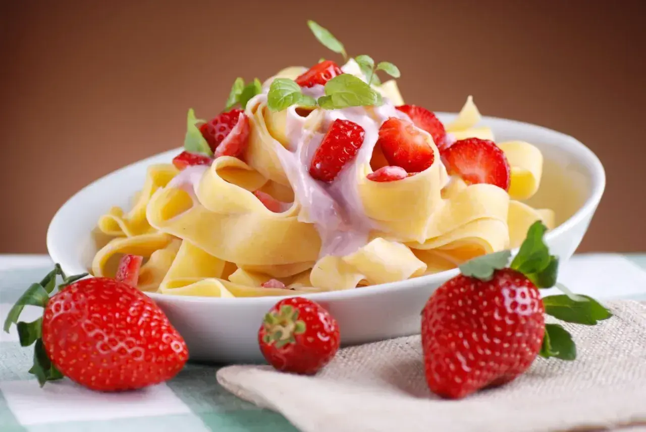 Pappardelle z kremowym sosem i kawałkami świeżych truskawek, ozdobione listkami mięty. Obok talerza leżą dojrzałe truskawki.