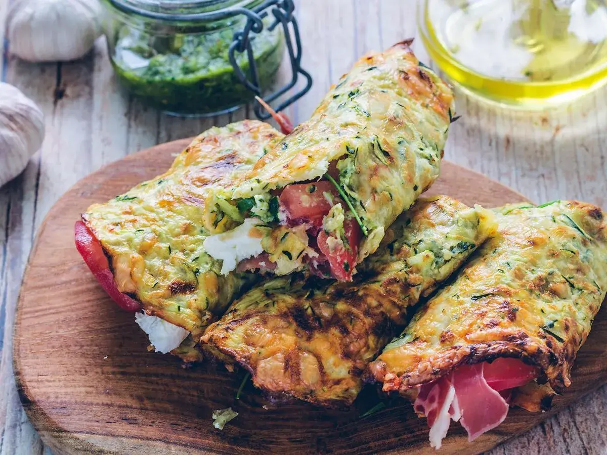 Złociste tortilla z cukinii, nadziewane pomidorami, fetą i szynką, prezentowane na drewnianej desce.