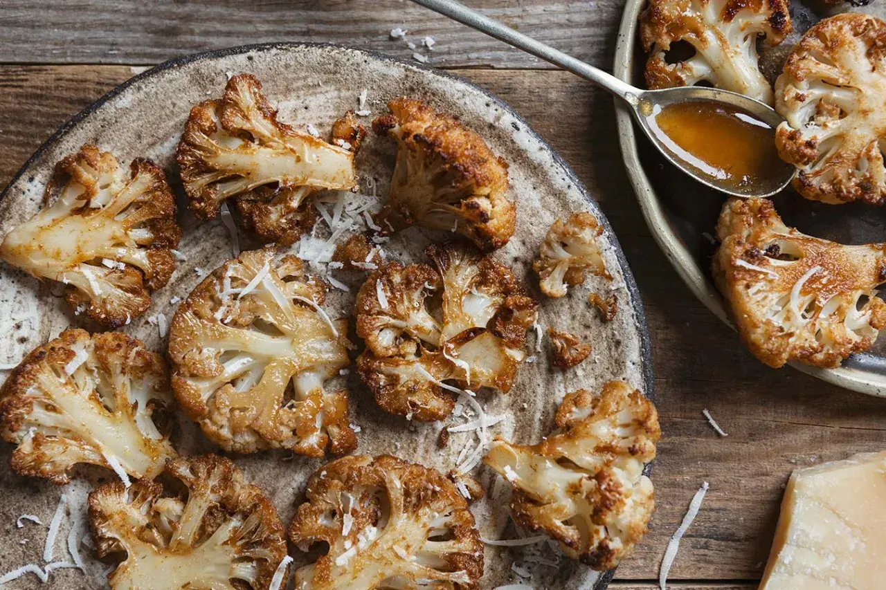 Pieczone dania z kalafiora, posypane startym parmezanem, na rustykalnym talerzu. Obok łyżka sosu i kawałek sera.
