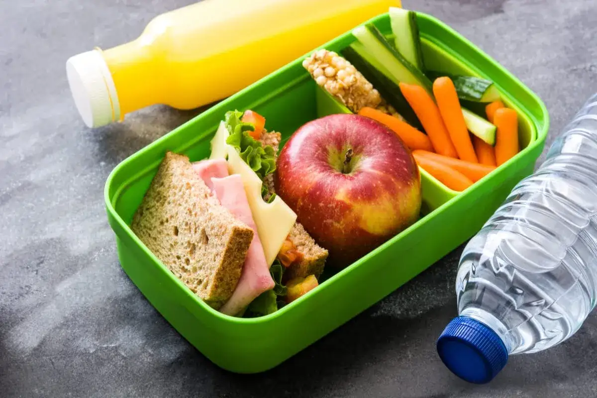Zielony lunchbox z kanapkami, jabłkiem, warzywami i batonikiem musli. Idealny pomysł na śniadanie w biegu. Obok butelka wody i soku.