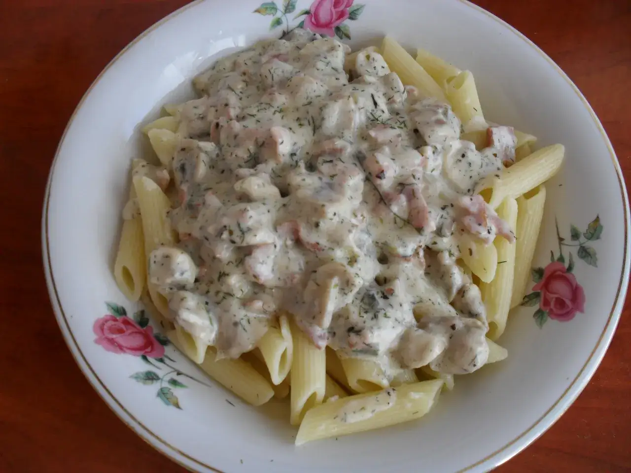 Penne z kremowym sosem, kawałkami pieczarek i boczku, posypane koperkiem. Pyszny makaron z pieczarkami na talerzu z różami.