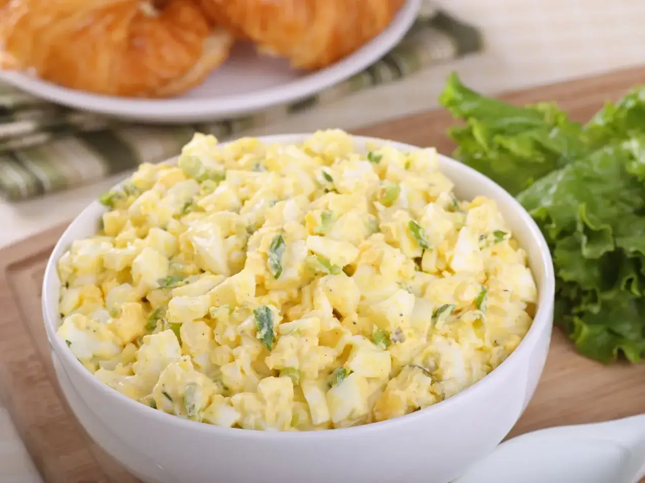 Pyszna sałatka z jajkiem, idealna na śniadanie lub lunch. Podana w białej miseczce, obok świeżej sałaty i chrupiących croissant&oacute;w.