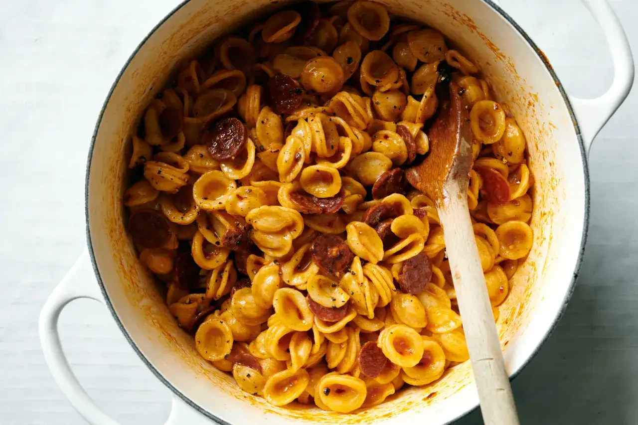 Orecchiette z pikantnym chorizo w sosie pomidorowym, mieszane drewnianą łyżką w białym garnku.