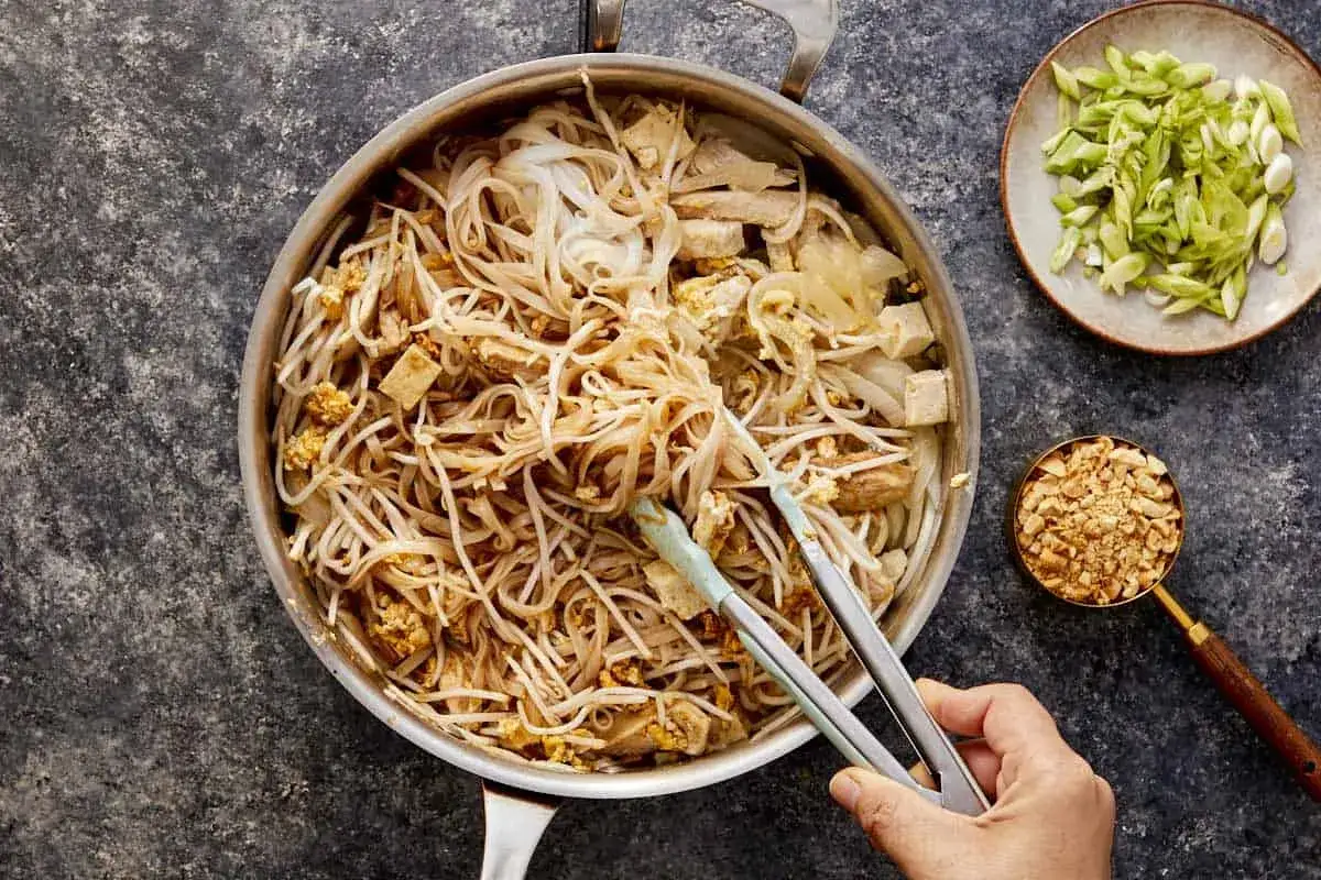 Gotowe do podania makaron po tajsku z kurczakiem i tofu, obok miseczka z orzeszkami ziemnymi i szczypiorkiem.