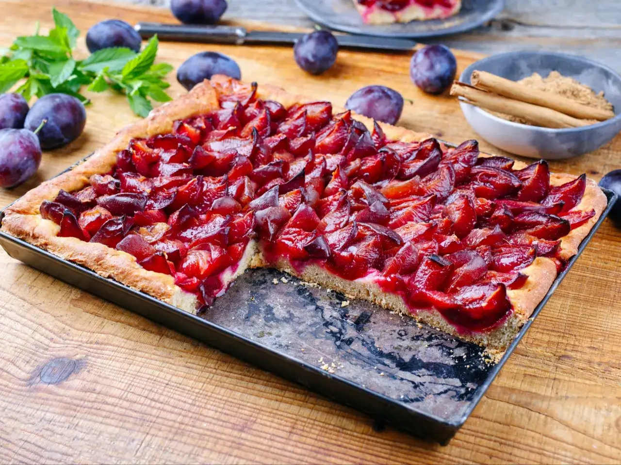 Pyszne ciasto ze śliwkami, świeżo upieczone, z rumianą skórką i soczystymi owocami. Obok leżą śliwki i laseczki cynamonu.