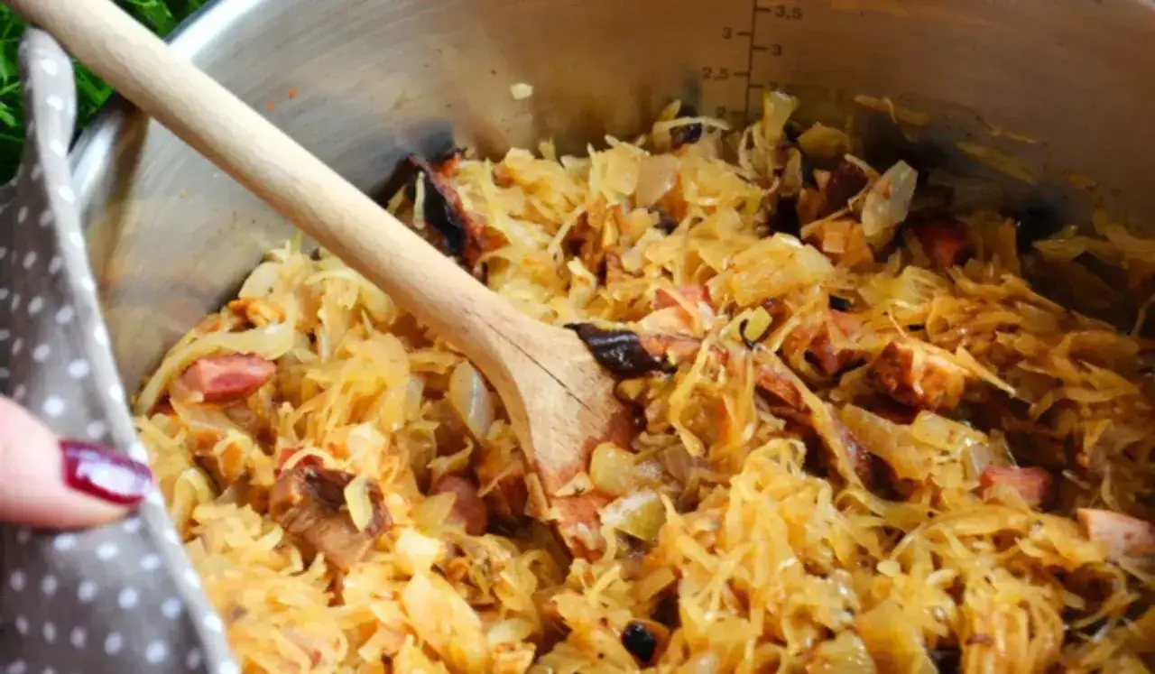Gotujesz bigos? Zobacz, jakie mięso do bigosu dodać, by był pyszny! W garnku kapusta z kawałkami wędzonego mięsa i kiełbasy, mieszana drewnianą łyżką.