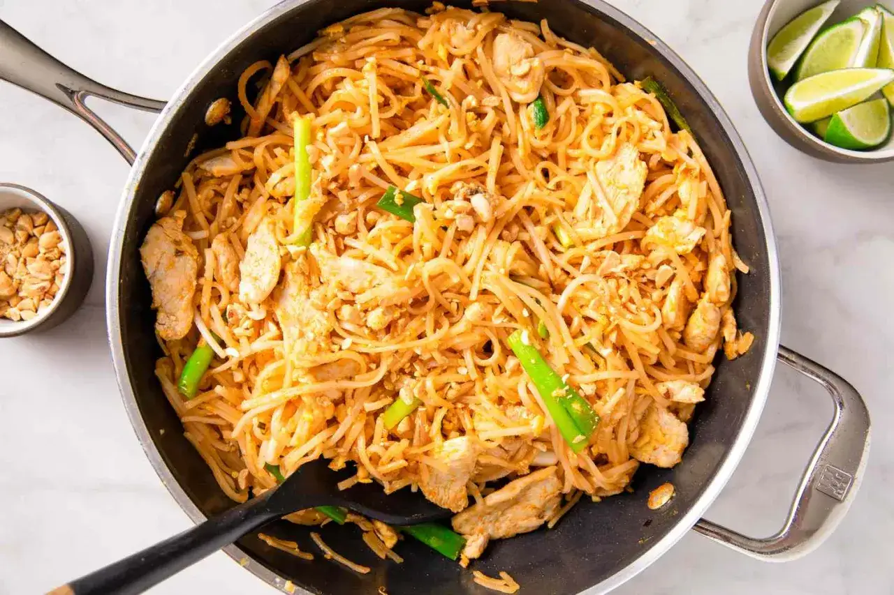 Gotowy do przygotowania pad thai przepis! Makaron ryżowy z kurczakiem, kiełkami i szczypiorkiem w woku. Obok miseczka z orzeszkami i ćwiartki limonki.