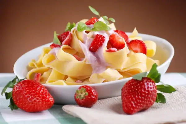 Pappardelle z kremowym sosem i kawałkami świeżych truskawek, ozdobione listkami mięty. Obok talerza leżą dojrzałe truskawki.