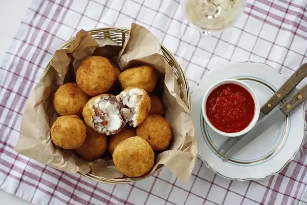 Koszyk pełen złocistych arancini, czyli smażonych kulek ryżowych z serem i mięsem. Obok miseczka sosu pomidorowego.
