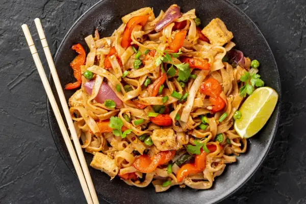 Pyszny pad thai przepis: makaron ryżowy z tofu, papryką, cebulą i groszkiem, udekorowany kolendrą i limonką.