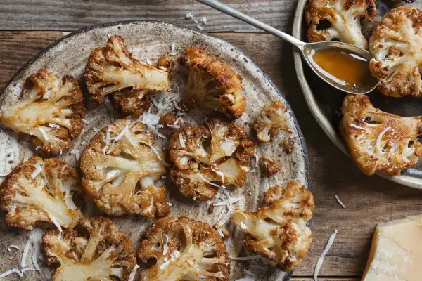 Pieczone dania z kalafiora, posypane startym parmezanem, na rustykalnym talerzu. Obok łyżka sosu i kawałek sera.
