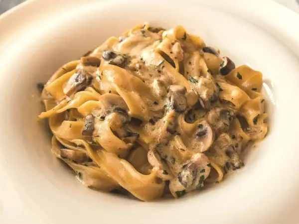 Pappardelle z kremowym sosem i kawałkami pieczarek, posypane natką pietruszki. Pyszny makaron z pieczarkami w białym talerzu.