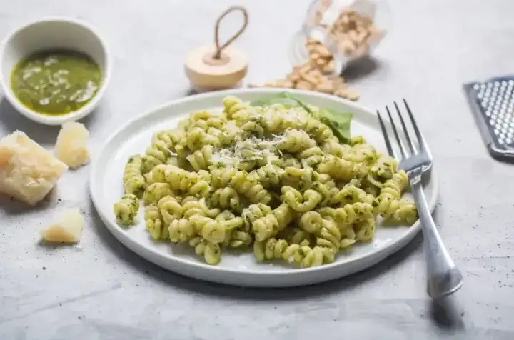 Pyszny makaron z pesto, posypany parmezanem, obok kawałki sera i miseczka sosu. Idealny posiłek dla miłośników włoskich smaków.